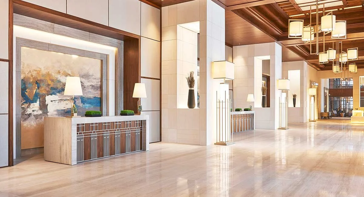 Elegant hotel lobby with modern decor, white and wood-paneled walls, large abstract artwork, multiple lamps, potted plants, and chandelier lighting.