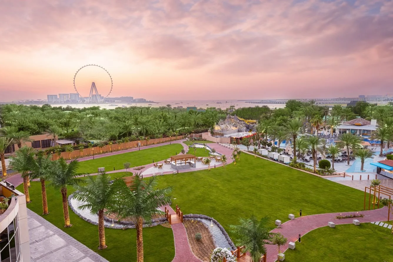 Sunset view over a lush green outdoor area with palm trees, pathways, a small bridge, and a building with pools and outdoor seating.