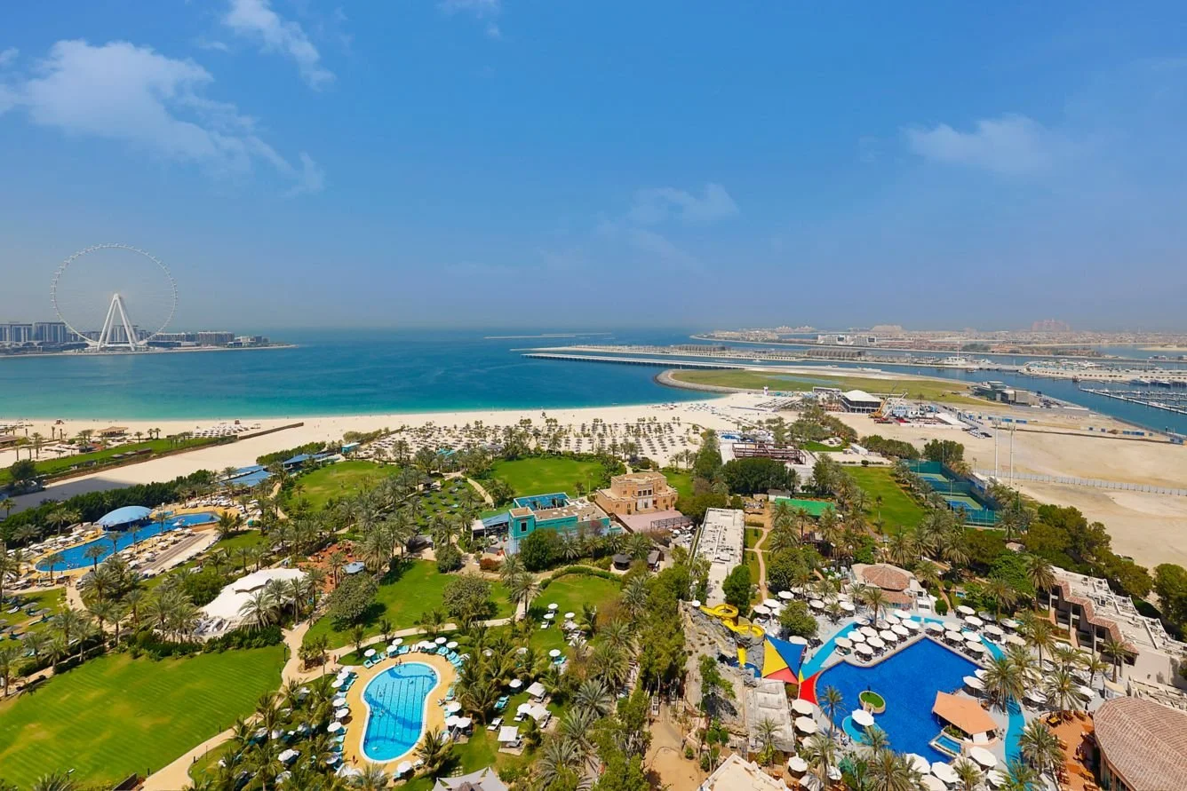 Aerial view of a coastal resort with pools, palm trees, a beach, and a Ferris wheel in the distance under a partly cloudy sky.