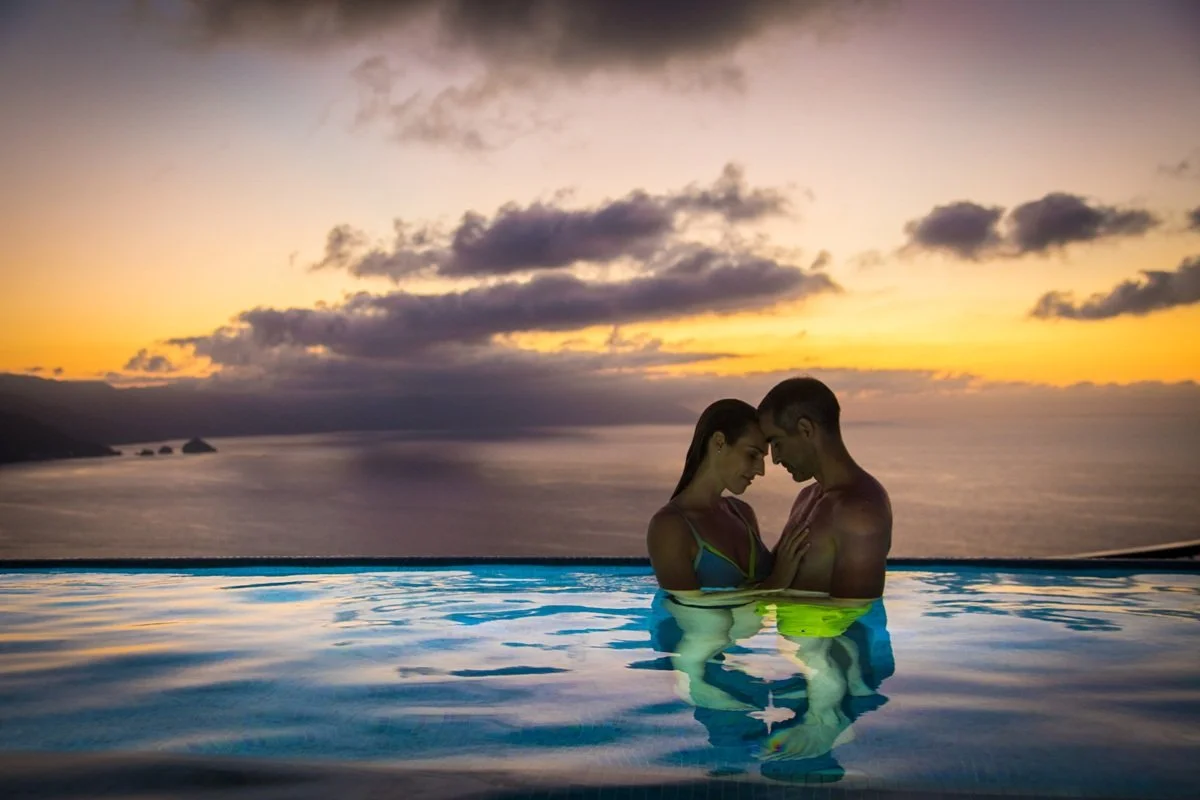A couple standing close together in a swimming pool at sunset, looking into each other's eyes with the ocean and a cloudy sky in the background.