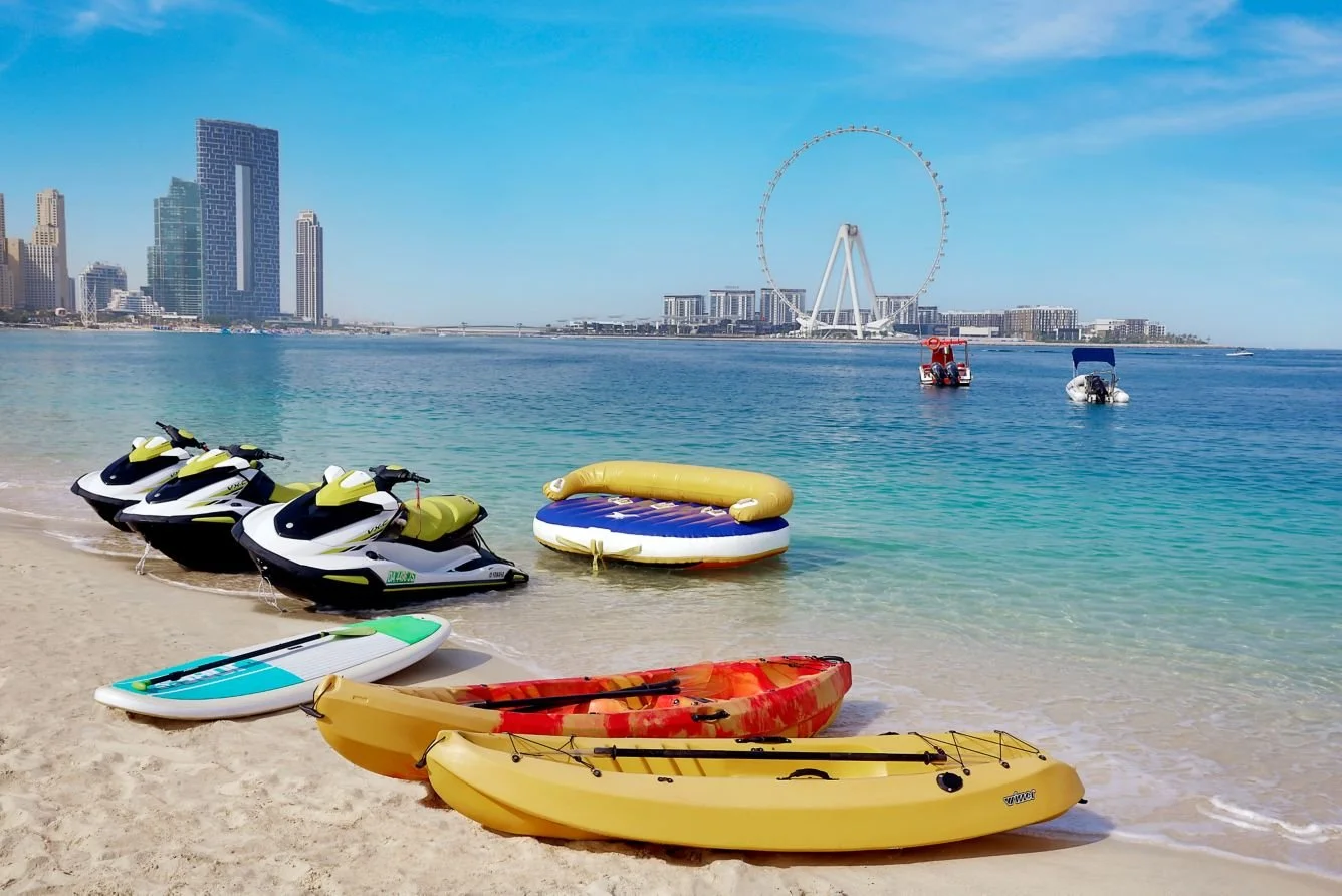Beach scene with jet skis, kayaks, and paddleboards on sandy shore, city skyline with skyscrapers, Ferris wheel, and calm water in background.