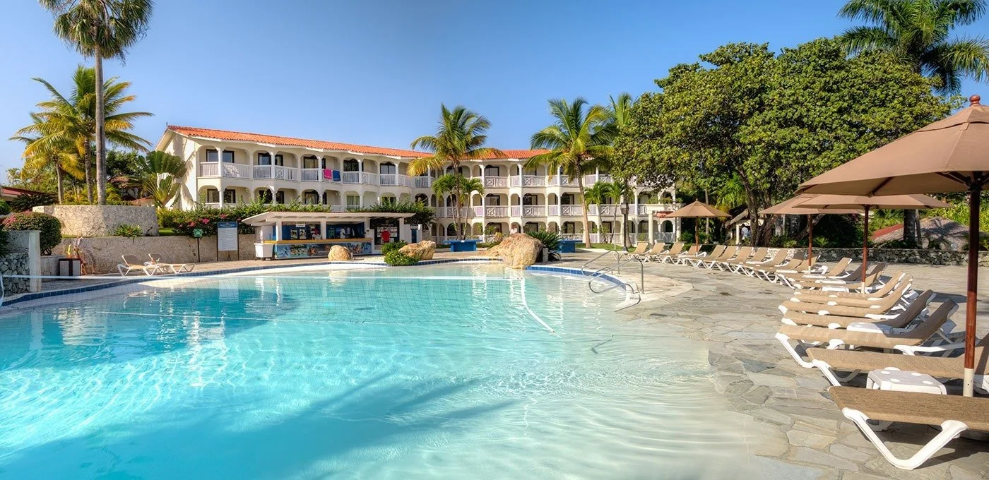 Hotel pool area with lounge chairs and umbrellas, multi-story hotel building in the background, palm trees, and clear blue sky, suggesting a tropical resort setting.