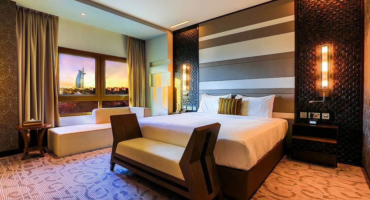 Modern hotel room with a large bed, a bench, a window seat, and cityscape view of the Burj Al Arab in Dubai at sunset.