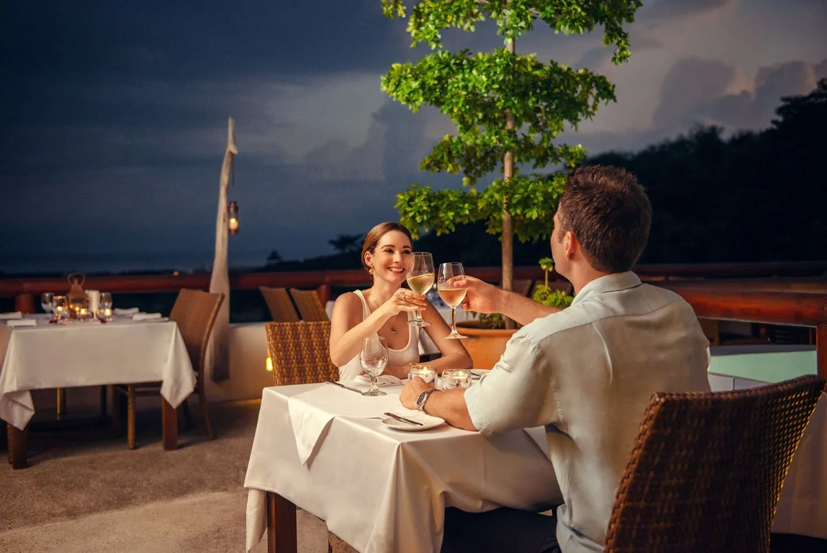 A couple enjoying dinner on an outdoor balcony at night, clinking glasses of white wine, with a lush green tree and a night sky in the background.