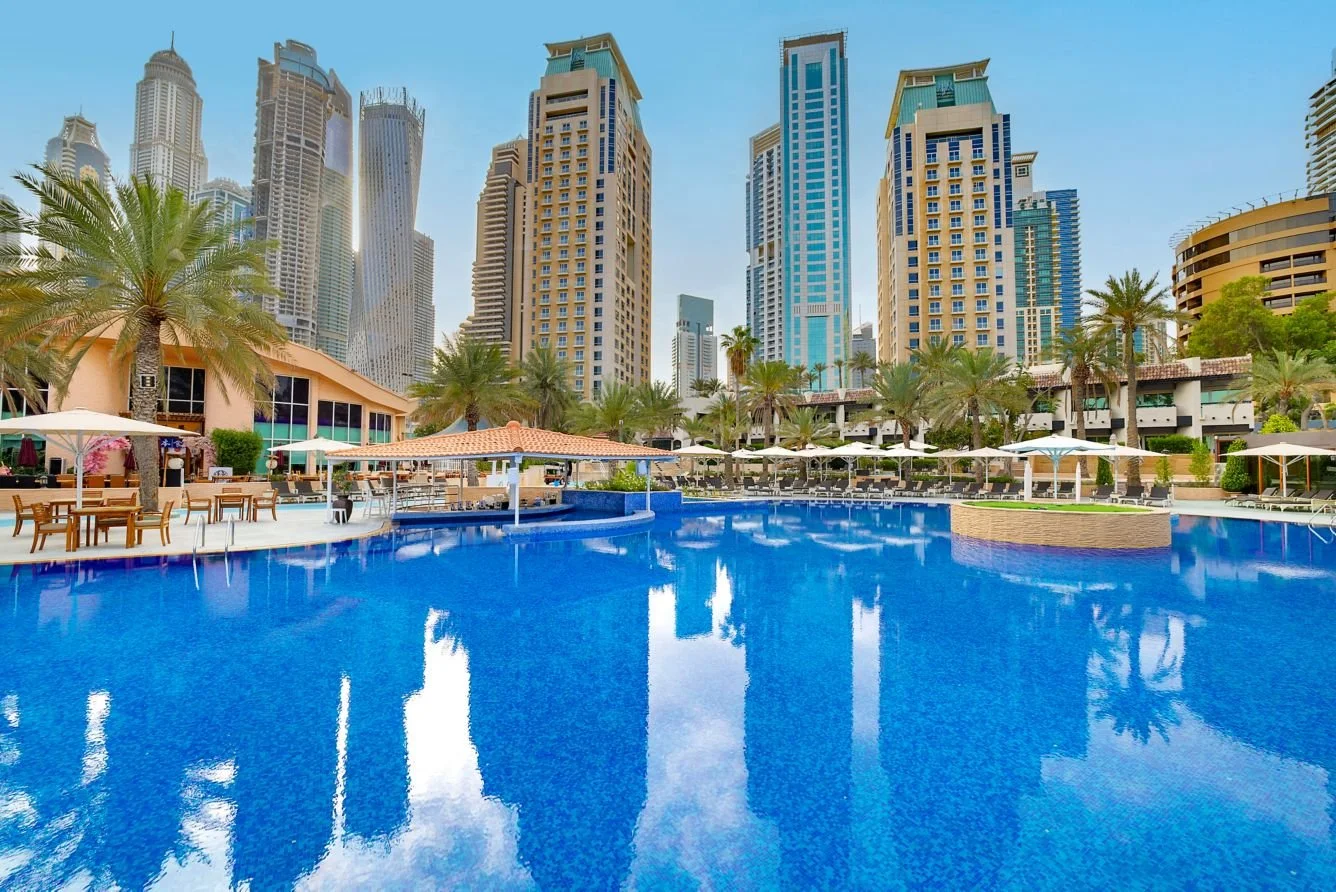 Poolside area at a hotel with luxury high-rise buildings in the background, surrounded by palm trees and sun loungers, under a clear blue sky.