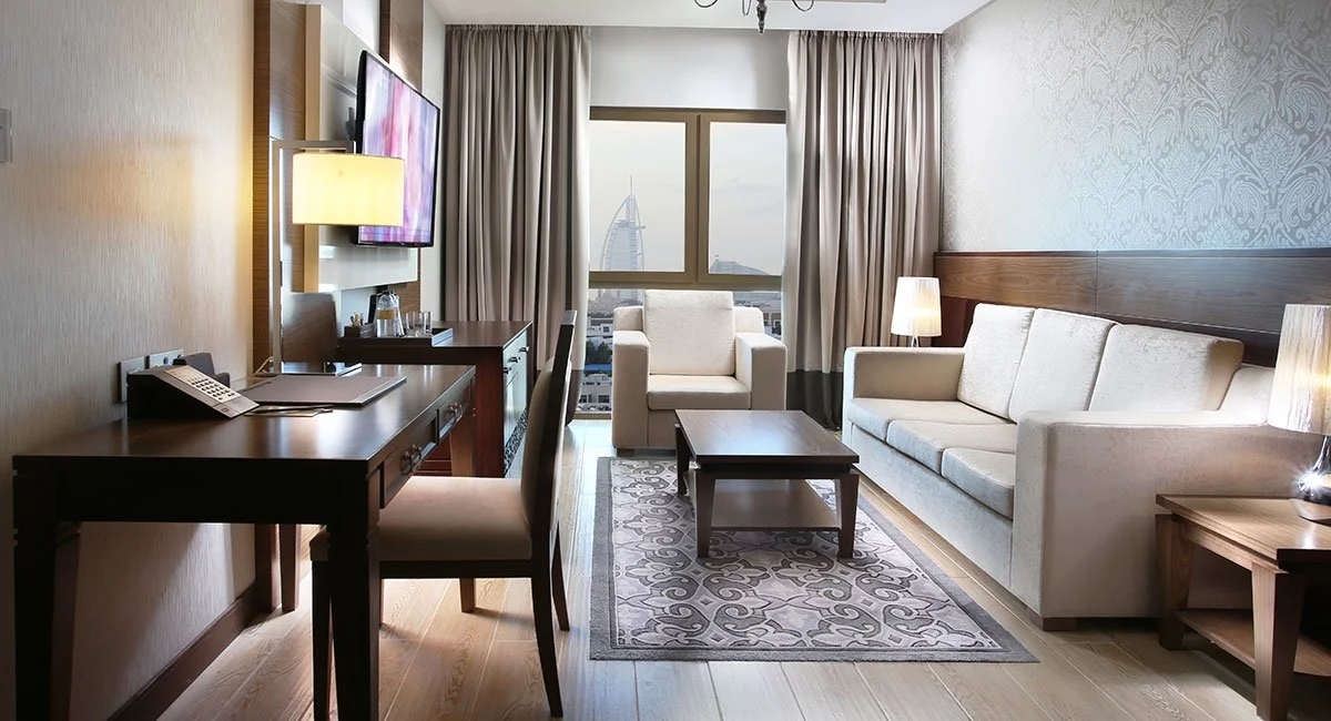 Hotel room with a white sofa, armchair, wooden coffee table, desk with a chair, wall TV, and a view of the Burj Al Arab hotel through the window.