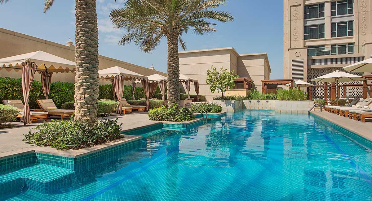 Swimming pool area with palm trees, lounge chairs under tents and umbrellas, modern buildings in the background, clear blue sky.