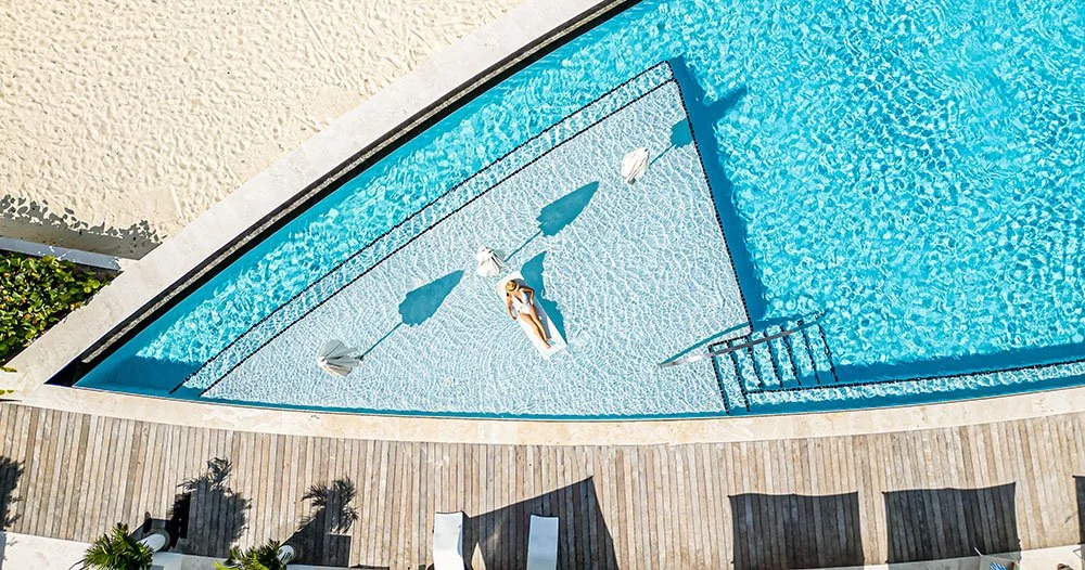 A woman relaxing on a float in a clear blue swimming pool, surrounded by lounge chairs, umbrellas, and a wooden deck, with stairs leading into the pool.