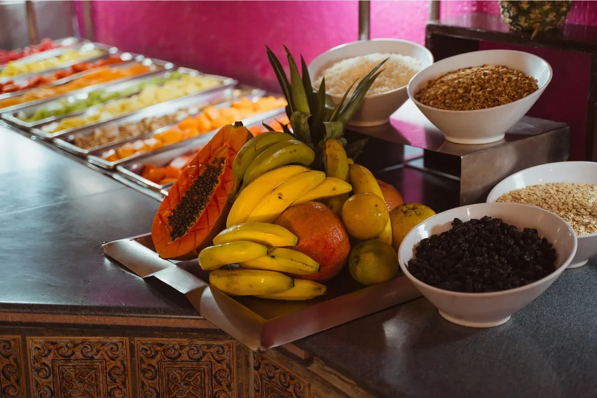 A tray of assorted tropical fruits including bananas, pineapple, an apple, and guavas, along with bowls of oats, granola, and dried fruits, set against a pink and dark background.