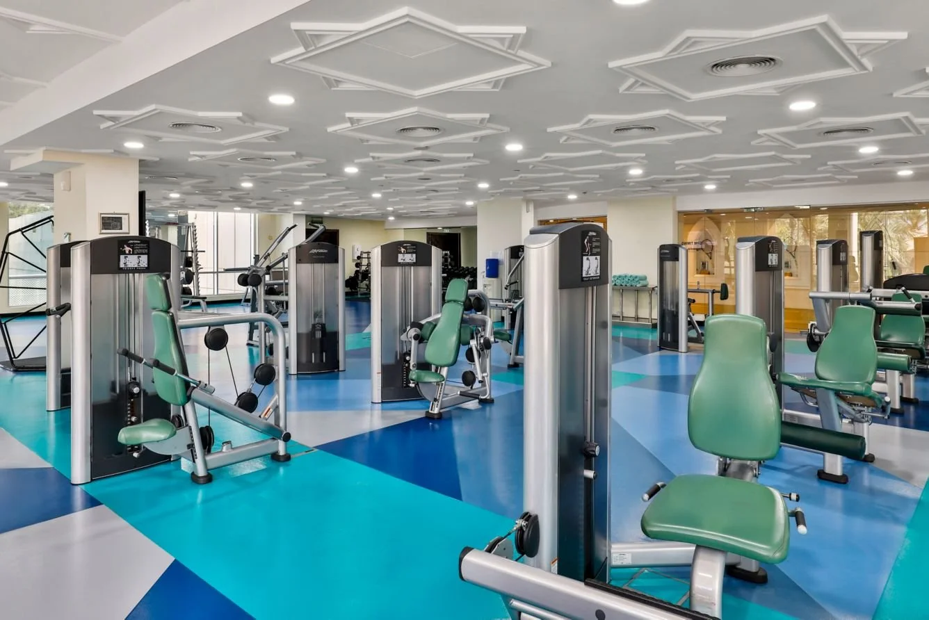 Empty gym with various exercise machines and strength training equipment, arranged in rows on a colorful floor with a geometric pattern, under a decorative ceiling with circular lighting.
