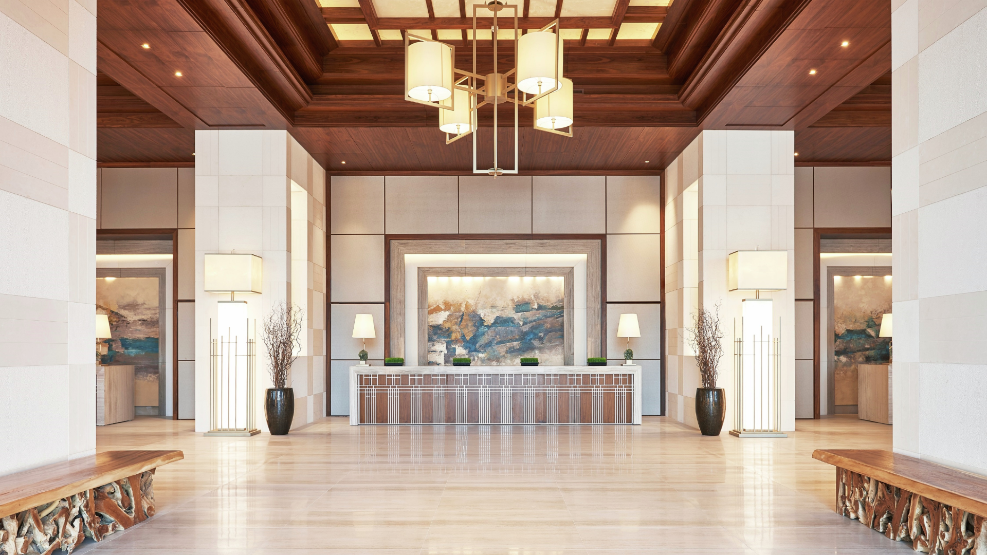 Elegant hotel lobby with a wooden reception desk, abstract artwork behind it, tall lamps, vases with decorative branches, and a wooden ceiling with a modern chandelier.