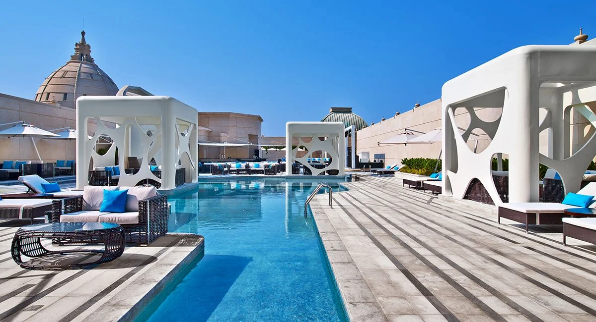 Luxury rooftop pool area with modern white structures, lounge chairs with blue cushions, and a cityscape in the background under clear blue skies.