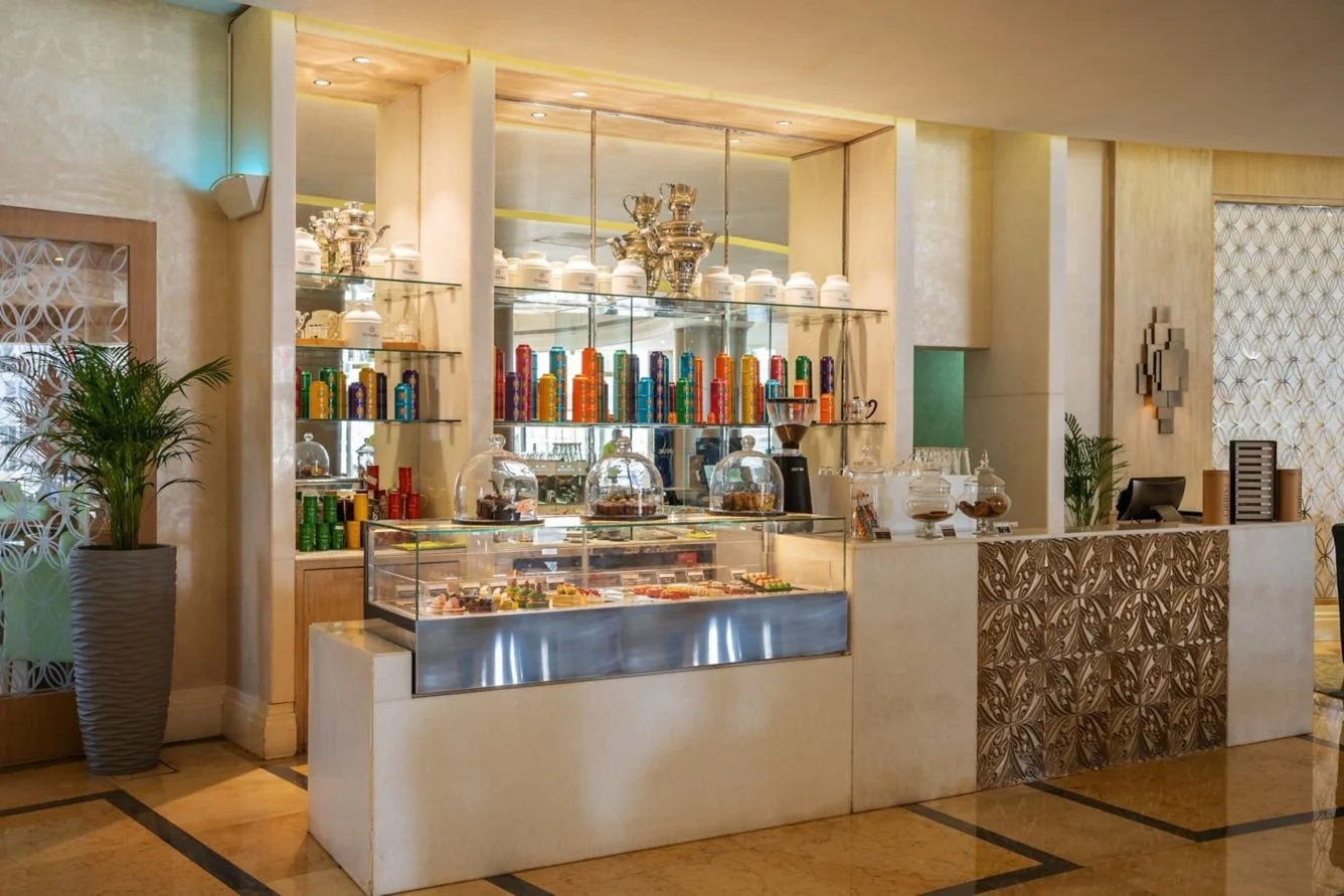 A dessert and coffee display at a cafe or hotel lobby with glass jars of cookies, colorful cans of soda, and a coffee machine, with a decorated front counter and potted plants on either side.