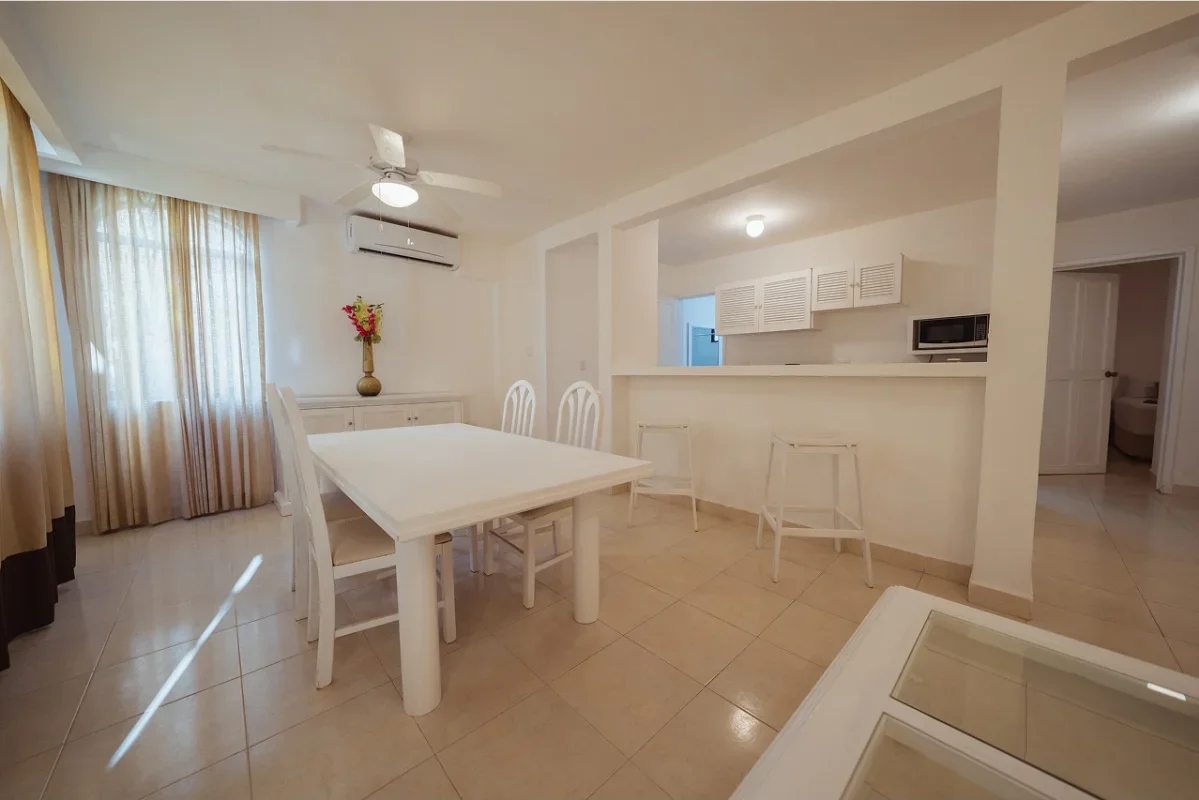 Dining area with a white table, four matching chairs, and a decorative vase with flowers on a sideboard. Curtains cover windows, and there is an air conditioner on the wall. The space is open to a kitchen and a hallway.