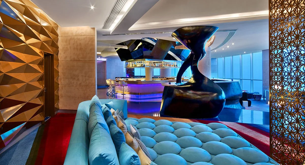 Luxurious modern hotel lobby features a curved blue sofa with decorative pillows, a striking abstract black sculpture, a sleek illuminated bar, and intricate geometric wood wall panels with large windows in the background.