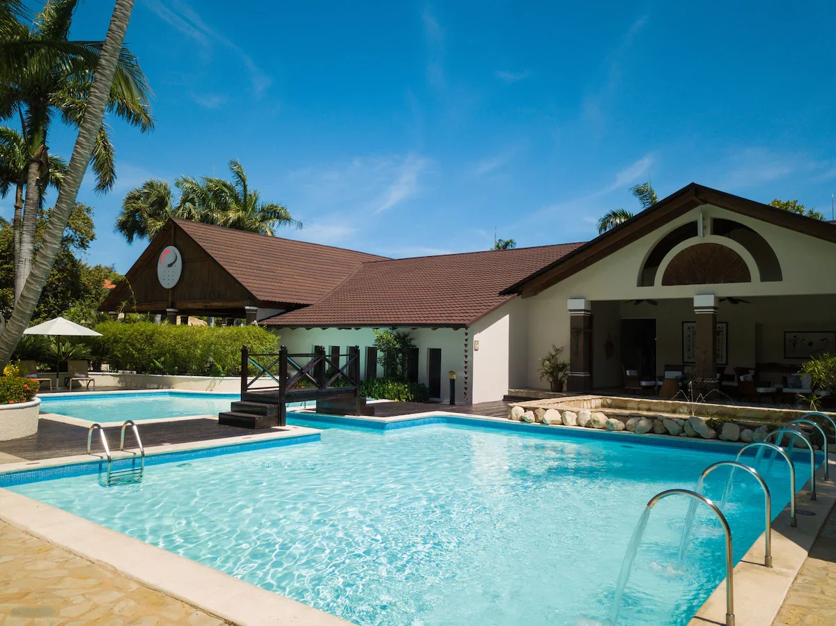 A luxurious outdoor swimming pool surrounded by palm trees, lounge chairs, and a poolside bar at a tropical resort with a building in the background and a clear blue sky.