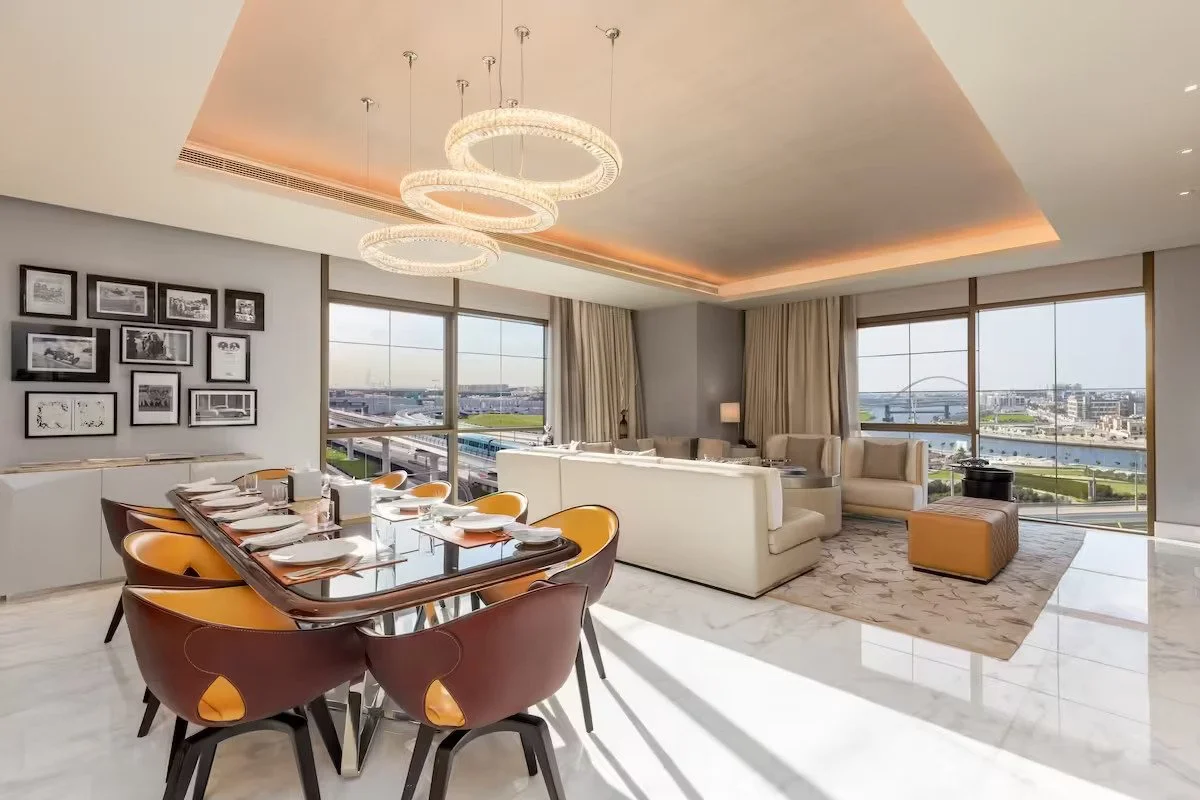 Modern dining and living room with large windows, chandelier, framed photos, dining table set, beige furniture, and city view