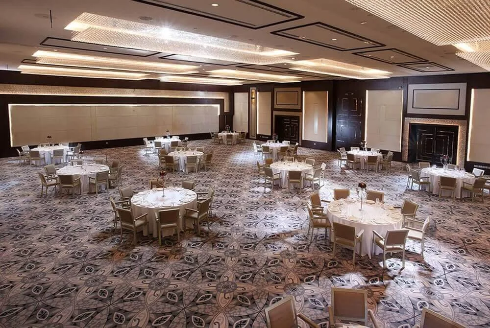 Empty banquet hall with round tables covered in white tablecloths and beige chairs, set for an event, with a patterned carpet and modern ceiling lighting.