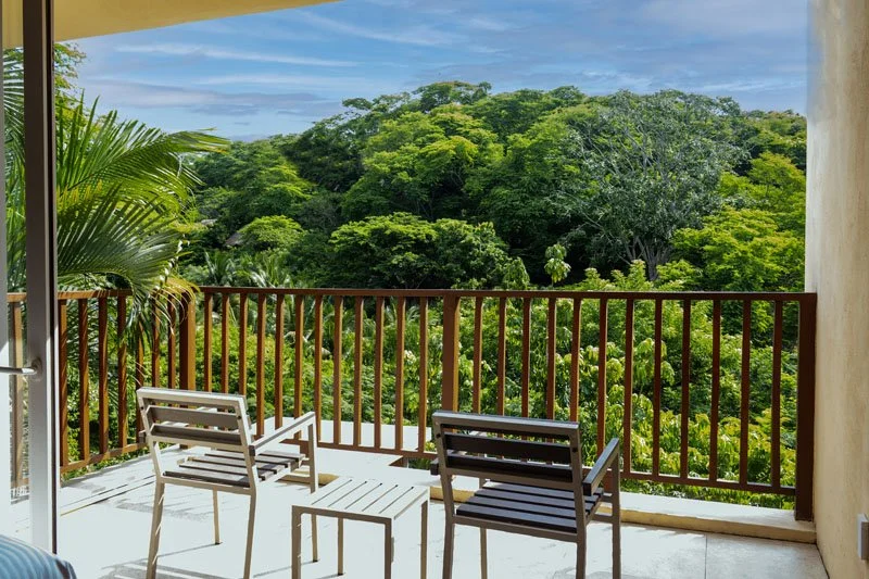 Balcony with two chairs and a small table overlooking a lush green forest under a partly cloudy sky.