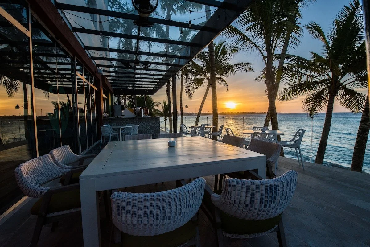 Outdoor dining area with white chairs and tables, overlooking the water with palm trees and a setting sun.