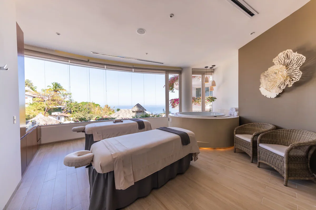 Spa room with massage tables near large window overlooking greenery and water, with seating area and decorative wall art.