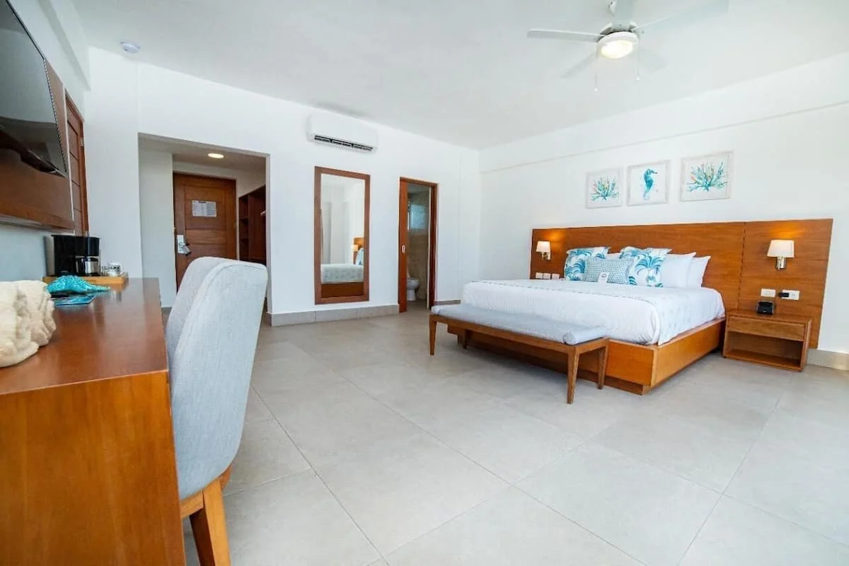 A hotel room with a large bed covered in white linens and decorative pillows, a wooden headboard with matching nightstands, wall-mounted lamps, a flat-screen TV, and a desk with a chair. There is a ceiling fan and a small bathroom visible in the back