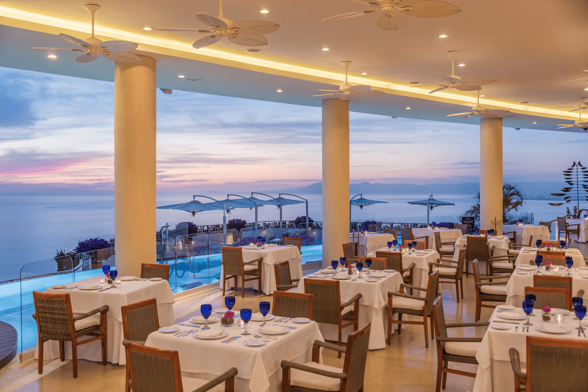 Elegant seaside restaurant with white tablecloths, blue glasses, and views of the ocean at sunset.