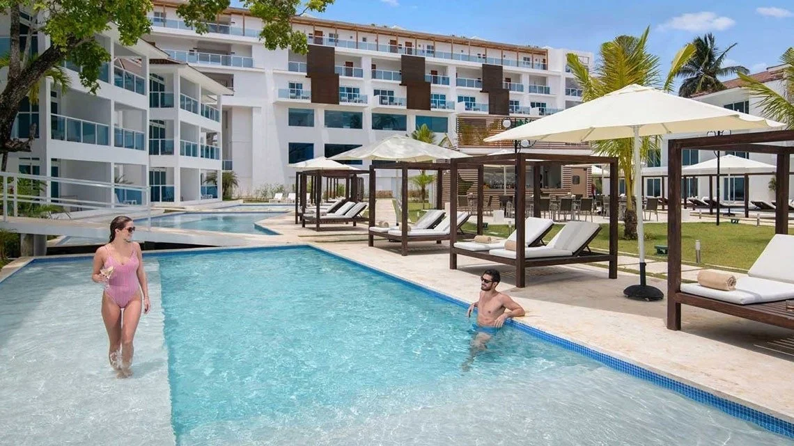 People enjoying a swimming pool at a resort, with lounge chairs and umbrellas nearby, modern apartment buildings in the background, and palm trees.