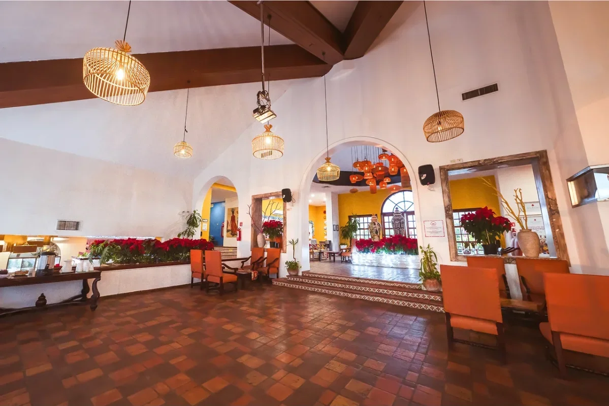 A cozy restaurant interior with terracotta tile flooring, white walls, and wooden ceiling beams. Wicker pendant lights hang from the ceiling. Plants and poinsettias decorate the space, which features an open area with a small raised platform and addi