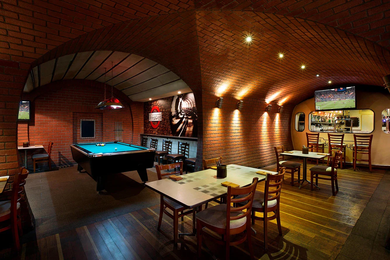 Interior of a bar or pub with a pool table, several tables and chairs, two TVs showing sports, brick walls, and warm lighting.