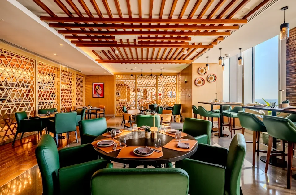 Elegant restaurant interior with round tables, green chairs, decorative wood paneling, and large windows letting in natural light.