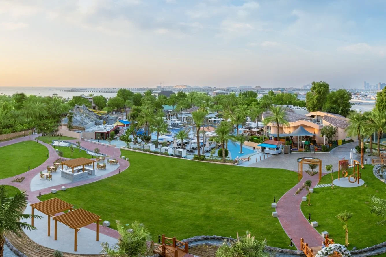A tropical resort with a swimming pool surrounded by palm trees, shaded seating areas, a green lawn, and pathways amid lush greenery, with cityscape and water in the background at sunset.