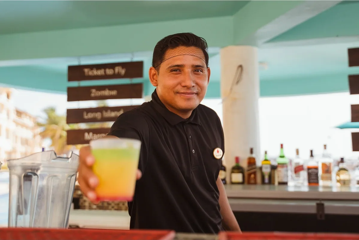 A smiling bartender in black shirt offering a colorful drink in a plastic cup in an outdoor setting with a bar, bottles, and signage in the background.