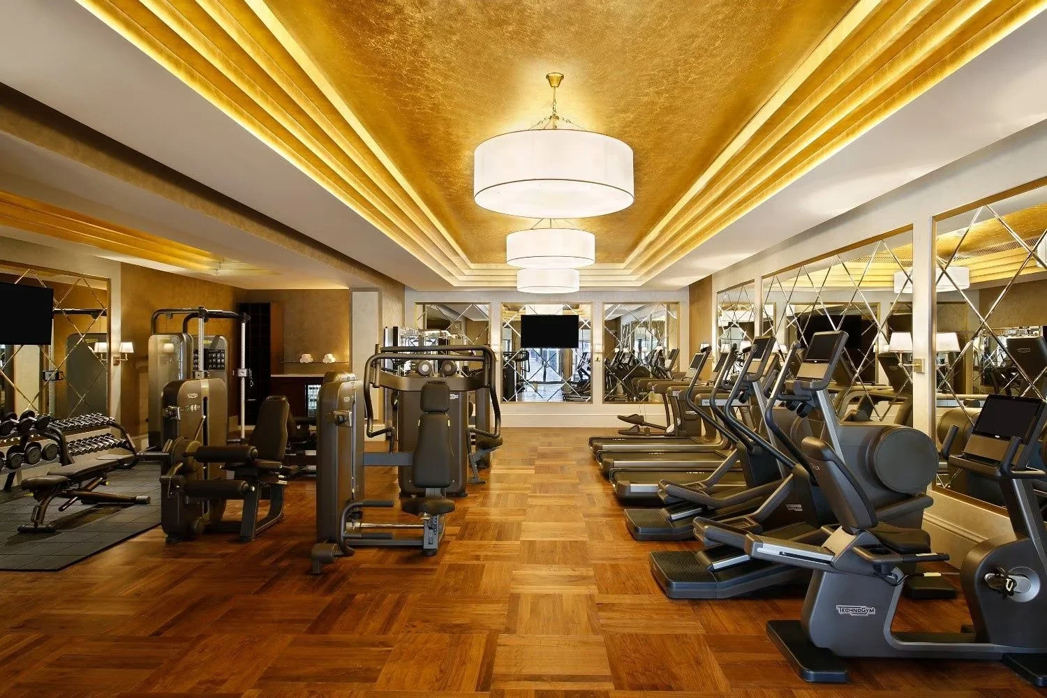 A luxurious gym with wooden flooring, mirrored walls, and ceiling lights. Equipped with treadmills, ellipticals, and weight machines, with gold and white decor.