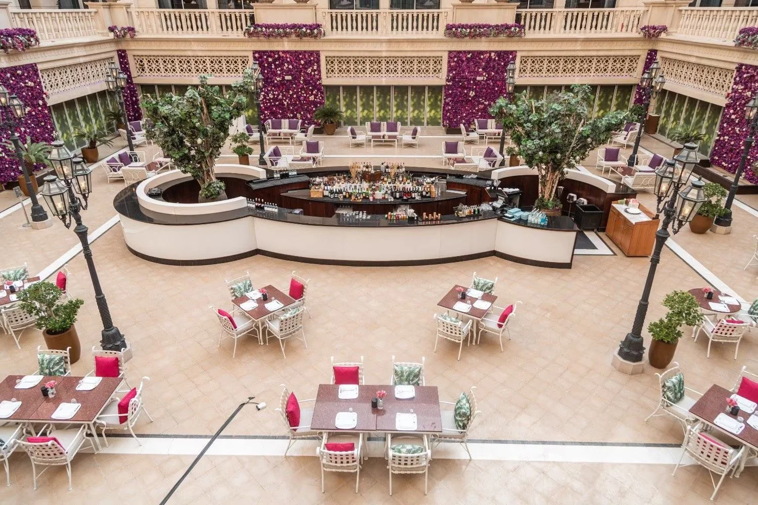 An elegant hotel lobby converted into a bar and dining area with a central curved bar, surrounded by tables with chairs, potted plants, and purple floral wall decorations.