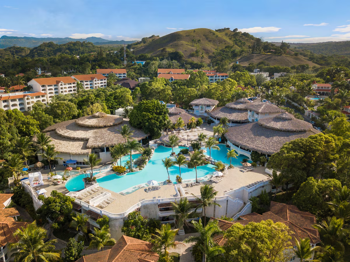 Aerial view of a tropical resort with multiple thatched-roof buildings, a large freeform swimming pool lined with palm trees, lounge chairs, and surrounded by lush green trees and hills in the background.