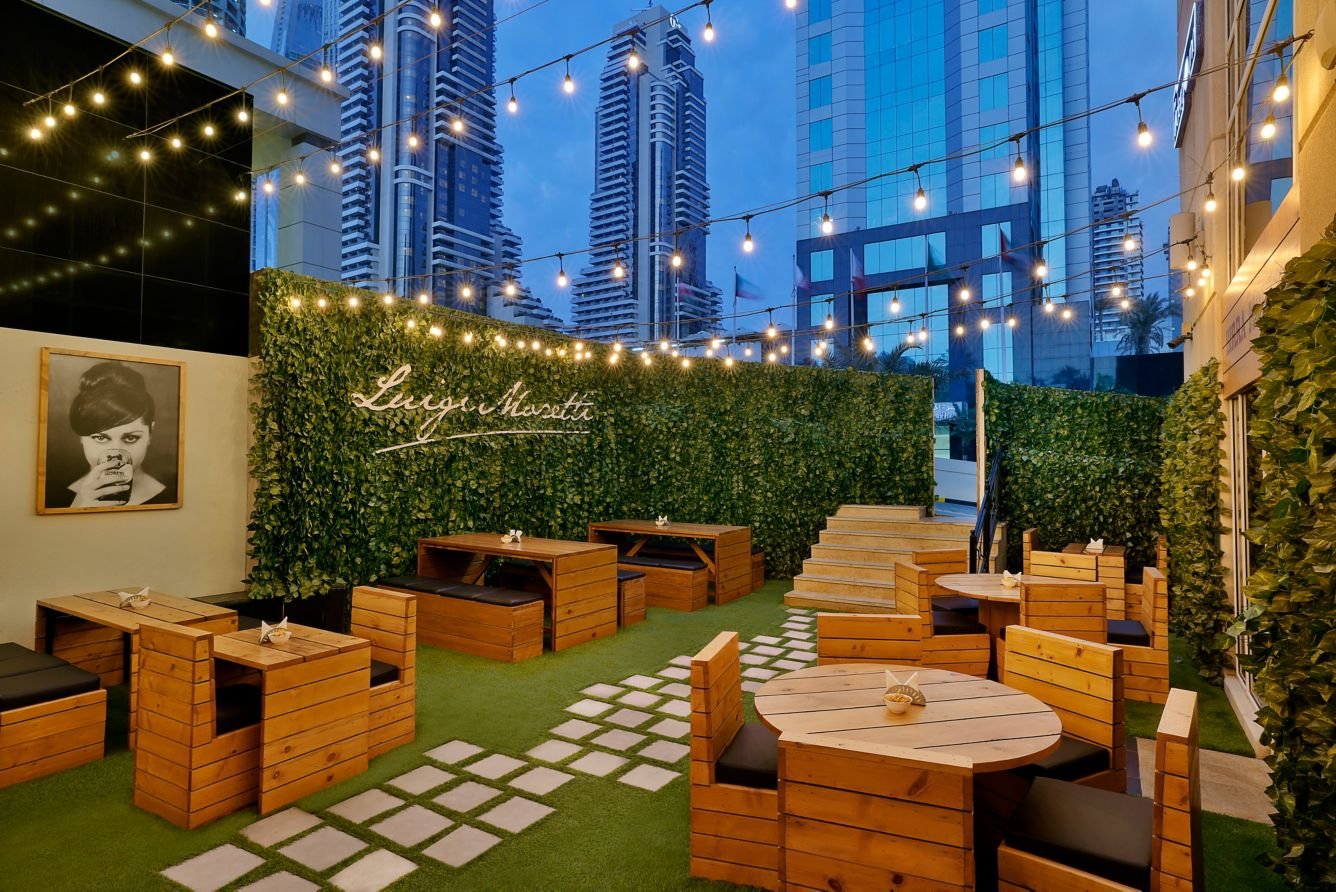 An outdoor restaurant patio with wooden tables and chairs, string lights overhead, a green wall with the sign 'Lugi & Margeti', and high-rise buildings in the background at dusk.