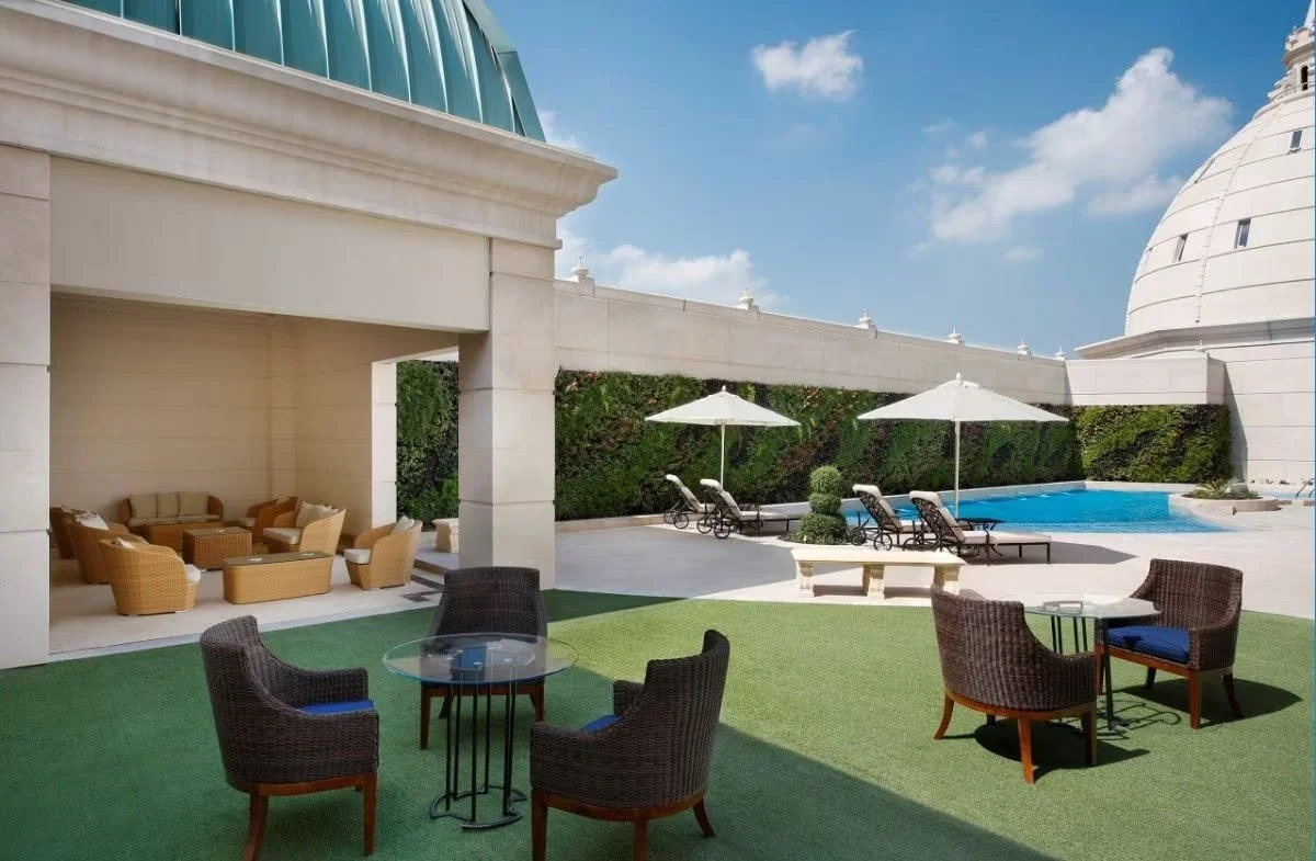 Luxury outdoor pool area with lounge chairs, umbrellas, and a seating area on green artificial turf, with a modern building and blue sky in the background.