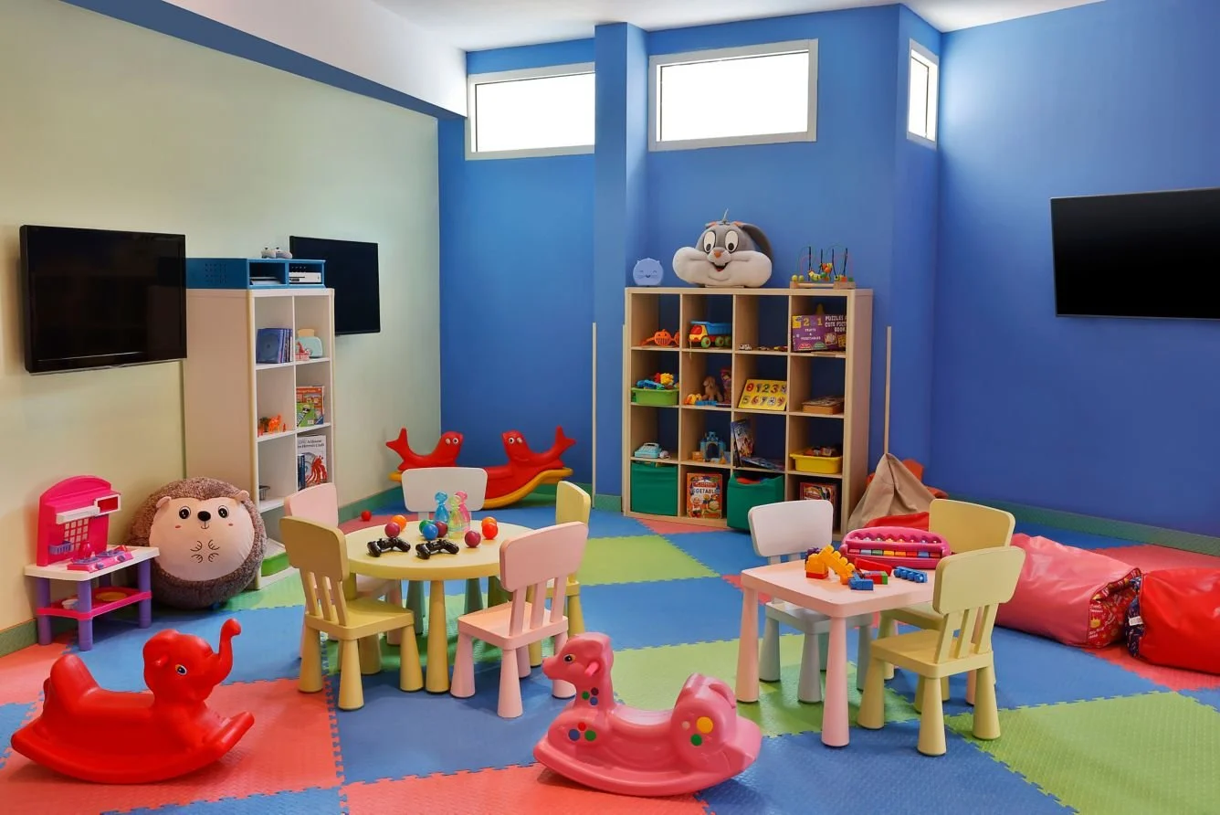Colorful children's playroom with toys, small tables and chairs, foam mats, and a large plush toy on a shelf. The room has blue and green walls, with two mounted televisions.