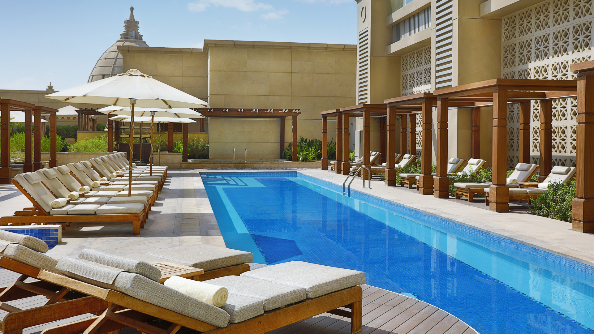 Resort swimming pool area with lounge chairs, umbrellas, and shaded cabanas, surrounded by a modern building and greenery.