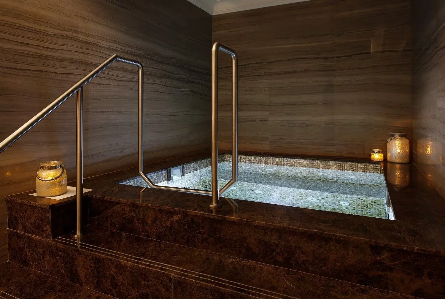 Indoor hot tub with tiled interior, surrounded by dark marble ledge and brown wooden walls, with candles atop jars placed at the corners for ambiance.