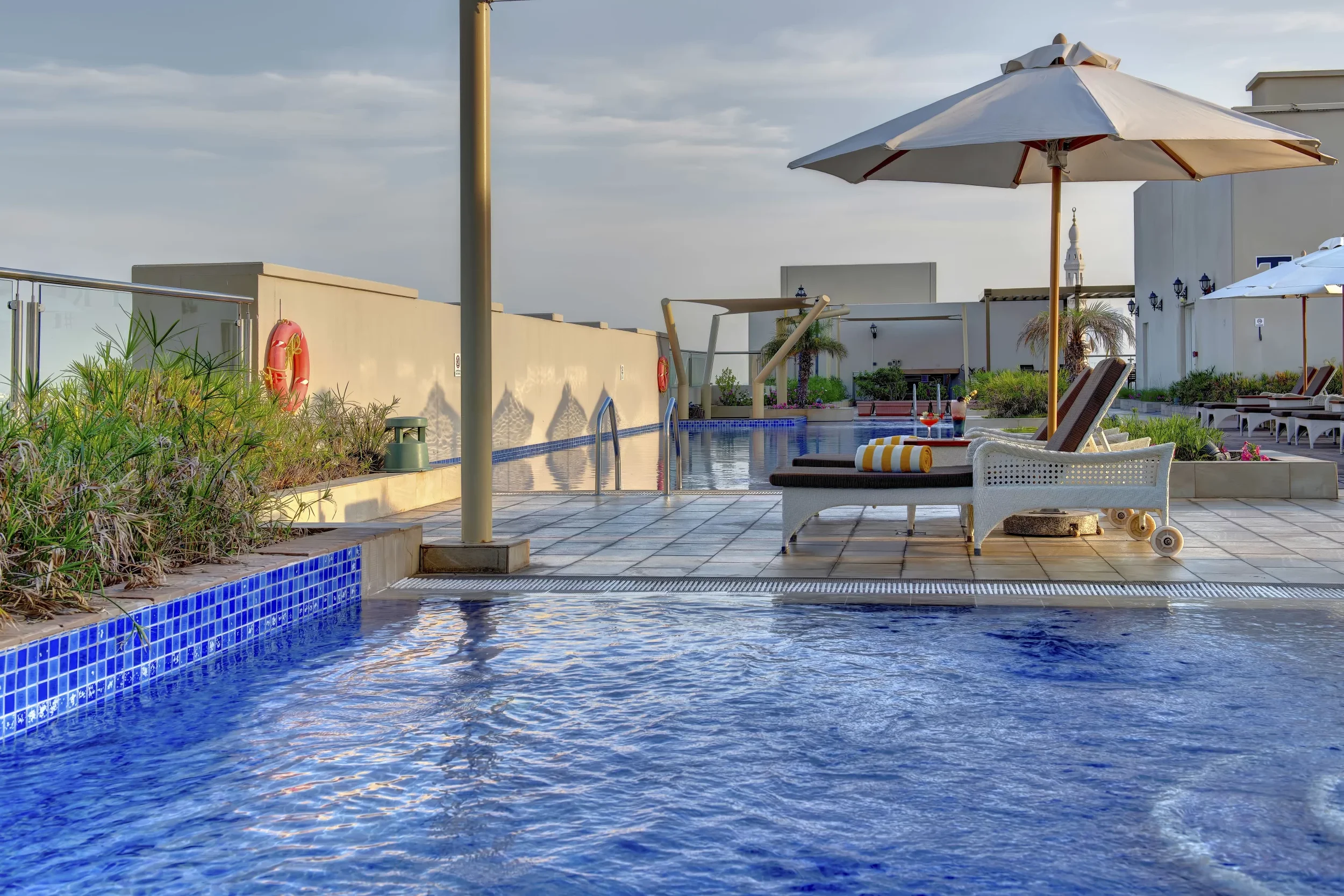 A rooftop swimming pool area with lounge chairs, large white umbrellas, and surrounding plants, under a partly cloudy sky.