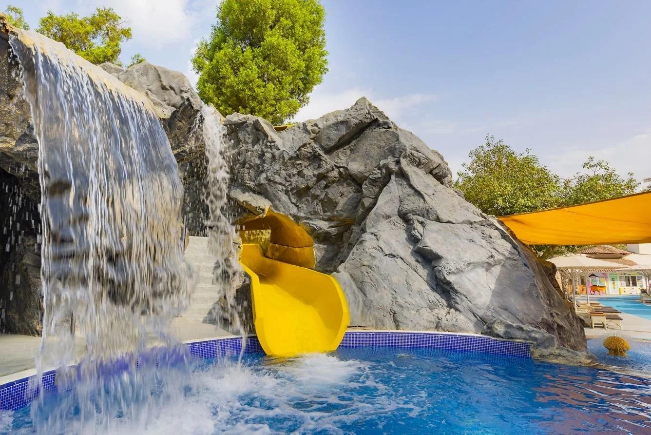 A water slide features a bright yellow slide with a waterfall and rocks at an outdoor water park, with trees and blue sky in the background.