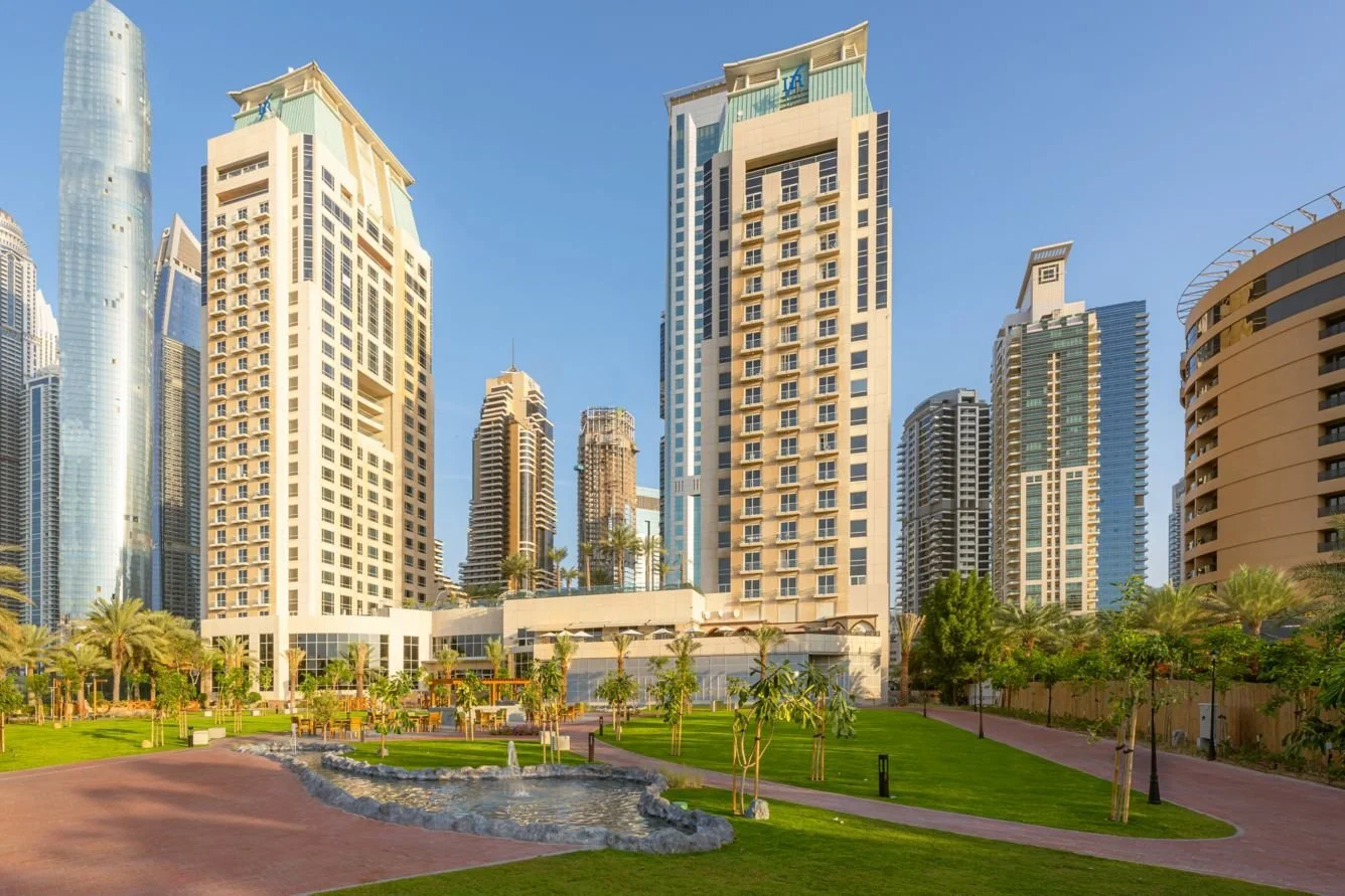 Sunlit modern cityscape with tall skyscrapers and a landscaped park featuring a small pond, palm trees, and walking paths.