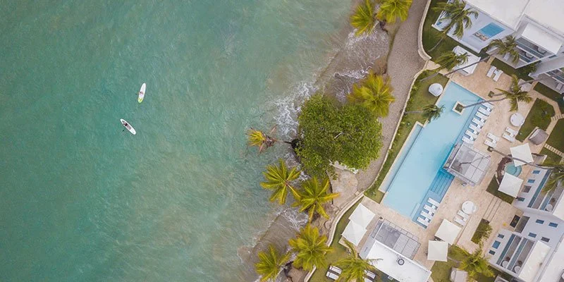 An aerial view of a beachfront hotel with a pool, surrounded by palm trees, sandy beach, and lounge chairs, with two people paddleboarding in the water.