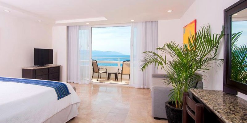 A hotel room with a bed, a flat-screen TV, a balcony with ocean view, a small table with two chairs, a large green plant, and a second TV on a granite counter.