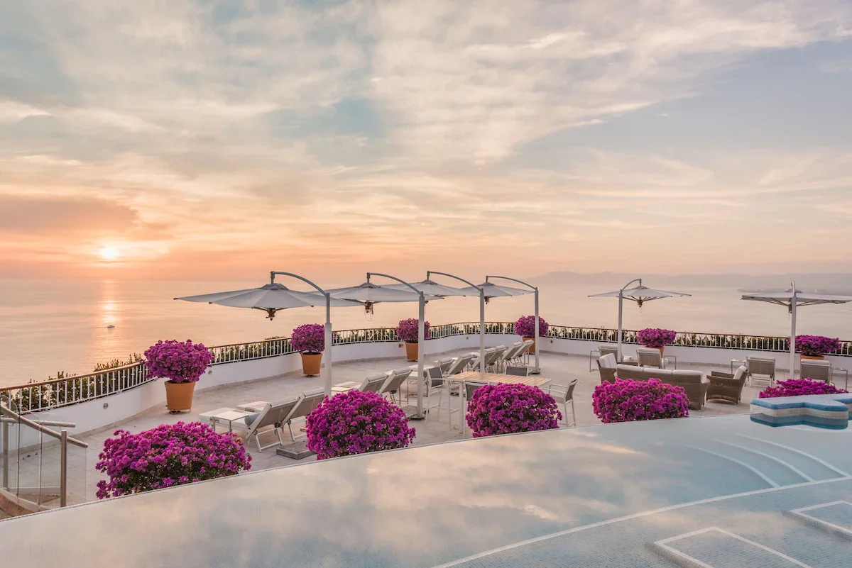 An outdoor terrace with pink flowers in pots, lounge chairs, and umbrellas overlooking the ocean during sunset.