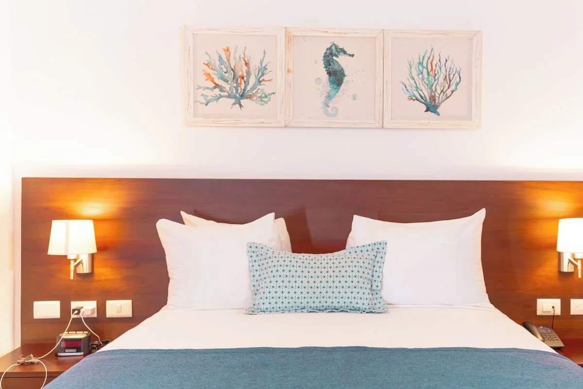 A neatly made hotel bed with white pillows, a decorative pillow, and a blue bedspread in a room with coral-themed artwork above the headboard and warm lighting from wall sconces.