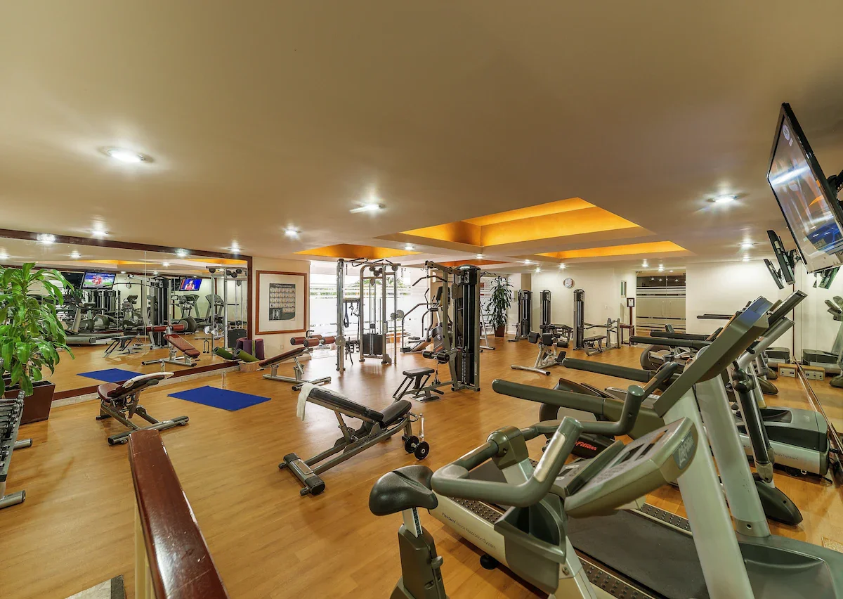 A spacious indoor gym with various exercise equipment, including treadmills, ellipticals, weight machines, and benches, with large windows allowing natural light.