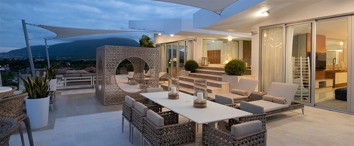 Outdoor rooftop terrace with dining area, lounge chairs, potted plants, and mountain view in the background at dusk.